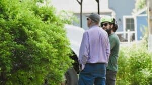 An ISA Certified Arborist in a green safety helmet and ear protection talks with a homeowner beside a work truck during an on-site tree assessment.
